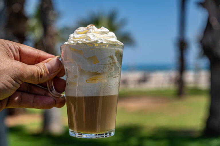 Mano sosteniendo un cafÃ© con nata en vaso transparente, fondo de palmeras y mar en un dÃ­a soleado.