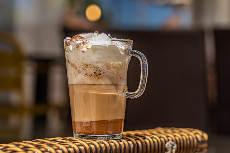 Taza de cafÃ© con espuma y cacao en un vaso transparente sobre una mesa de mimbre en una cafeterÃ­a.