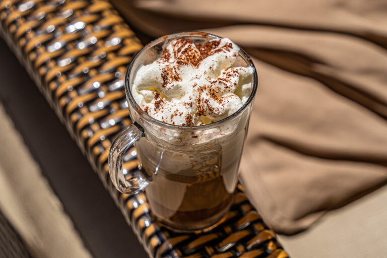Vaso de cafÃ© con nata montada y cacao en polvo sobre una mesa de mimbre en una terraza soleada.