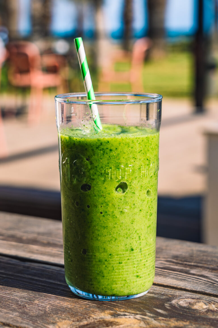 Batido verde en vaso alto con pajita a rayas verdes y blancas sobre mesa de madera en terraza al aire libre.