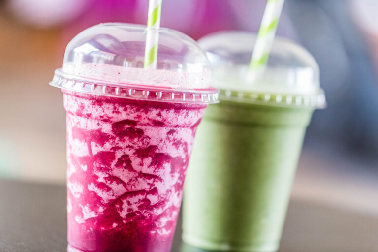 Batido rosa y batido verde en vasos de plÃ¡stico con tapas transparentes y pajitas sobre una mesa.