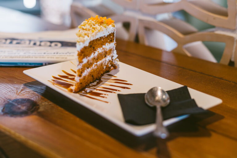 PorciÃ³n de pastel de zanahoria con crema y caramelo en plato blanco sobre mesa de madera, con cuchara al lado.