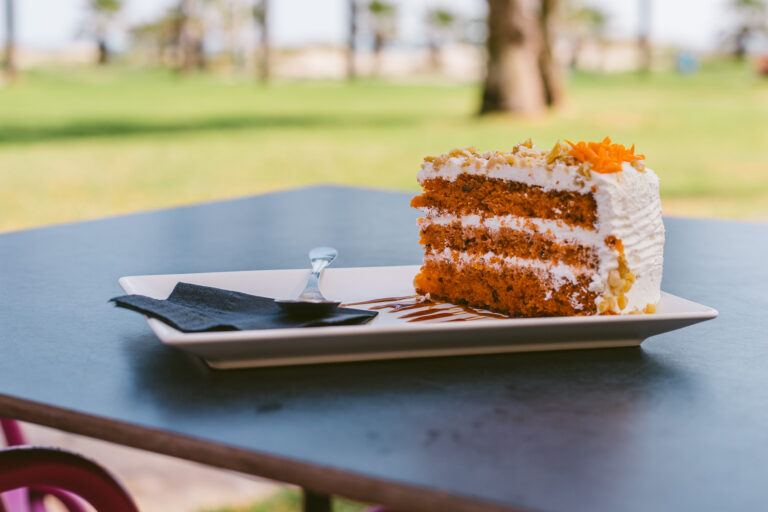 Trozo de pastel de zanahoria con crema en plato blanco sobre mesa al aire libre, fondo de cÃ©sped y palmeras.