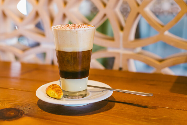 CafÃ© con leche condensada y espuma en vaso alto sobre plato con croissant pequeÃ±o, fondo decorativo en My Sweet CafeterÃ­a