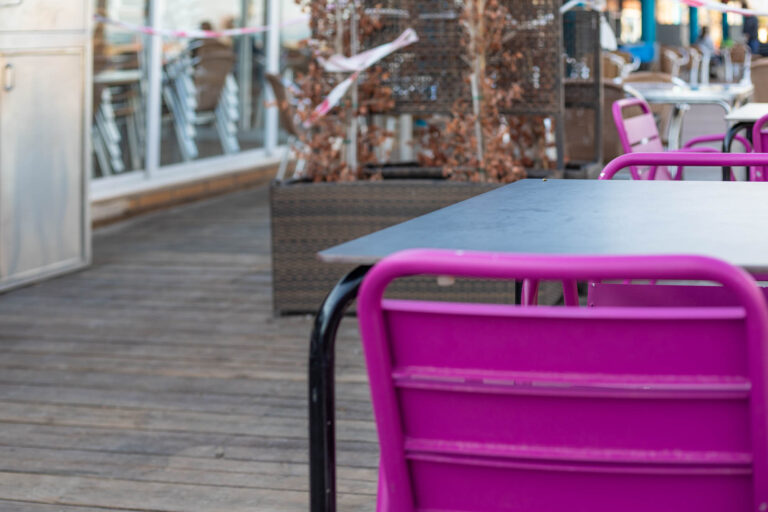 Terraza de cafeterÃ­a con sillas moradas y mesas negras sobre suelo de madera, plantas decorativas al fondo