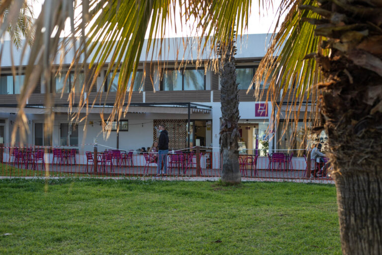 Terraza de cafeterÃ­a con mesas y sillas rosas, rodeada de palmeras y cÃ©sped, en un dÃ­a soleado.