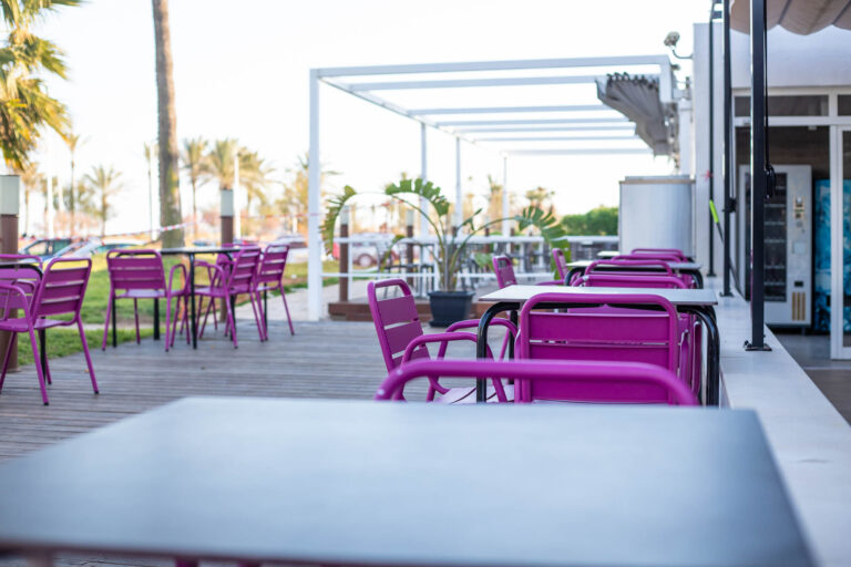Terraza de cafeterÃ­a con sillas moradas y mesas negras, rodeada de palmeras y estructura blanca en el Hotel Costa Azahar