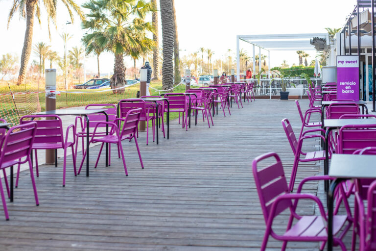 Terraza de cafeterÃ­a con sillas moradas y mesas en un paseo marÃ­timo, rodeada de palmeras.