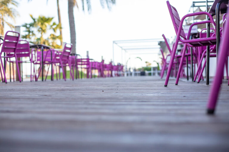 Terraza con sillas moradas y mesas vacÃ­as sobre suelo de madera, palmeras al fondo en un dÃ­a soleado.