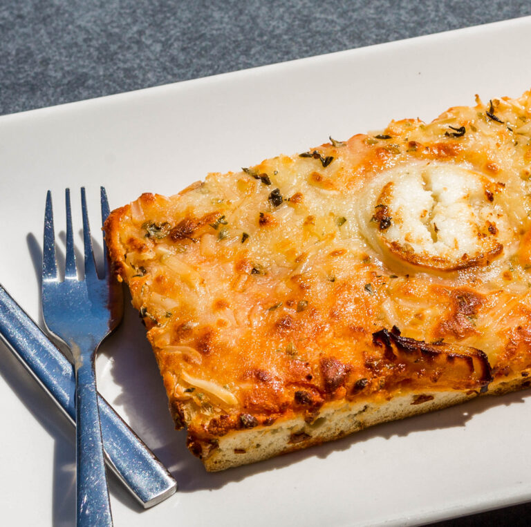 PorciÃ³n de pizza rectangular con queso derretido y cebolla sobre plato blanco, tenedor y cuchillo al lado.