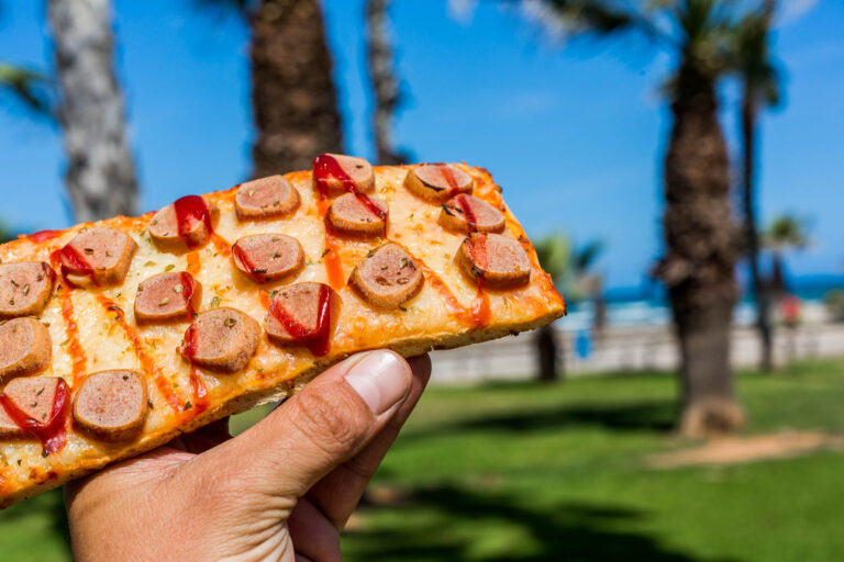 Pizza rectangular con salchichas y ketchup sostenida en mano, palmeras y cielo azul de fondo en Costa Azahar.